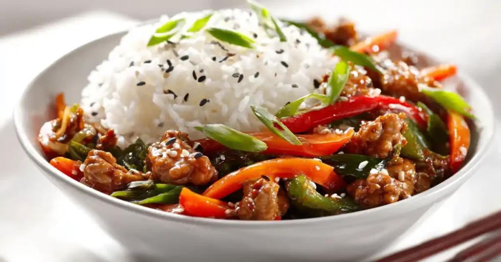 Ground chicken stir fry served with colorful vegetables and steamed jasmine rice in a white bowl topped with sesame seeds and green onions.